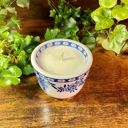 Candle in a decorative blue and white teacup on a wooden surface with green leaves in the background