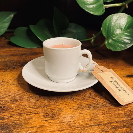 White mug with a lit candle on a wooden surface with green leaves in the background
