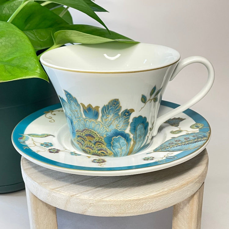 Teacup and saucer with floral design on a small wooden stool next to a plant.