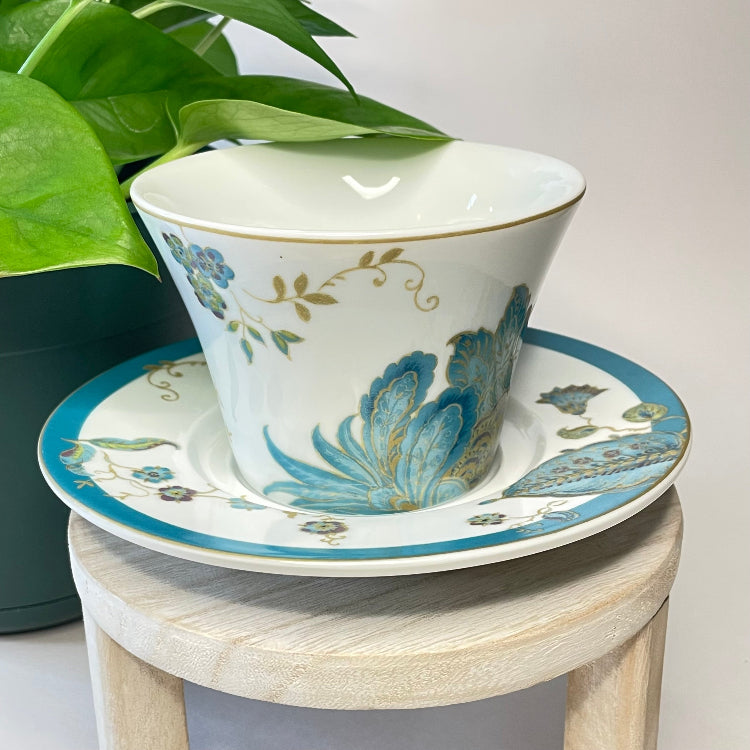 Ceramic cup and saucer with floral design on a small wooden stool, next to a potted plant.