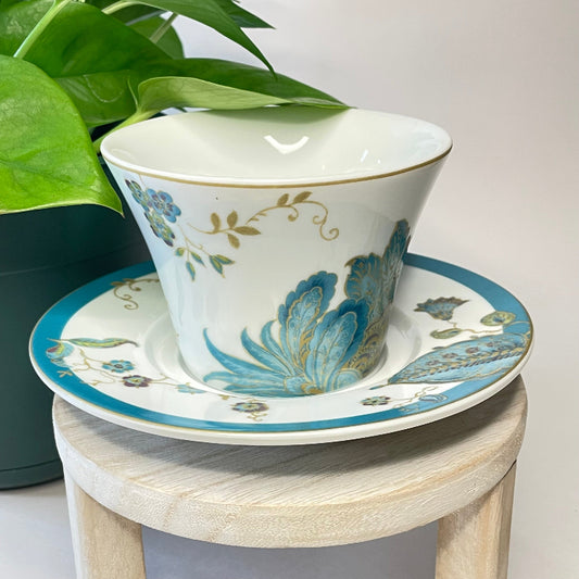 Ceramic cup and saucer with floral design on a small wooden stool, next to a potted plant.