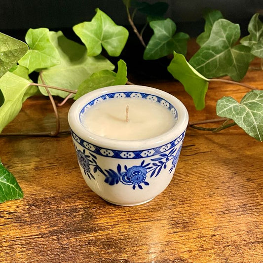 Candle in a blue and white ceramic teacup on a wooden surface with green leaves in the background