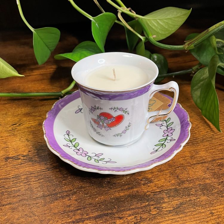 Candle in a decorative teacup on a matching saucer with green leaves in the background