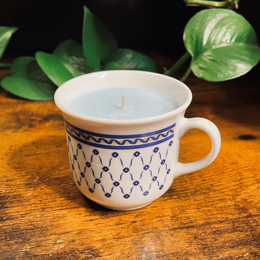 Candle in a white mug with blue patterns on a wooden surface with green leaves in the background