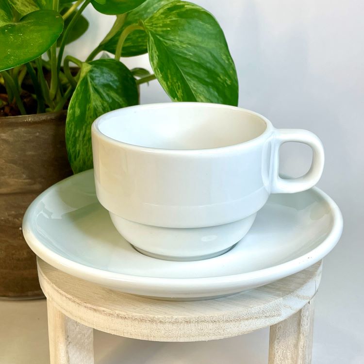 White ceramic cup and saucer on a small wooden stool with a plant in the background