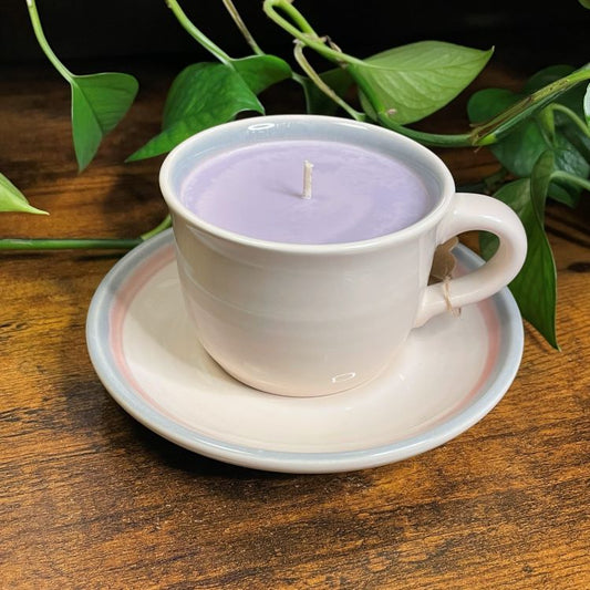 Candle in a white cup on a saucer with green leaves in the background