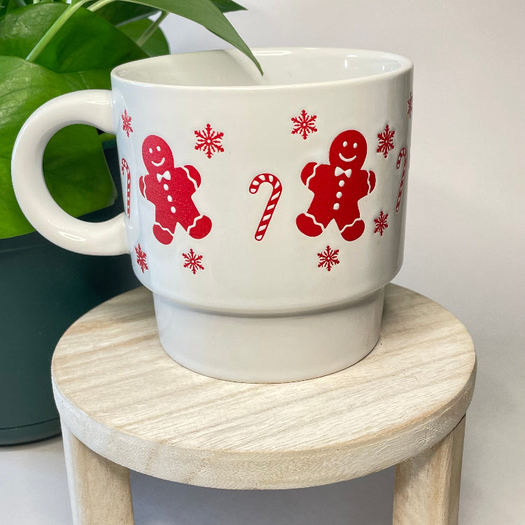 White mug with red gingerbread man and candy cane design on a wooden stool.