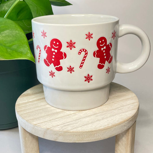 White mug with red gingerbread men, candy canes, and snowflakes on a wooden stool.