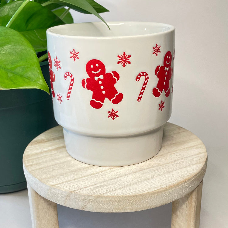 White pot with red gingerbread man and candy cane design on a wooden stool.