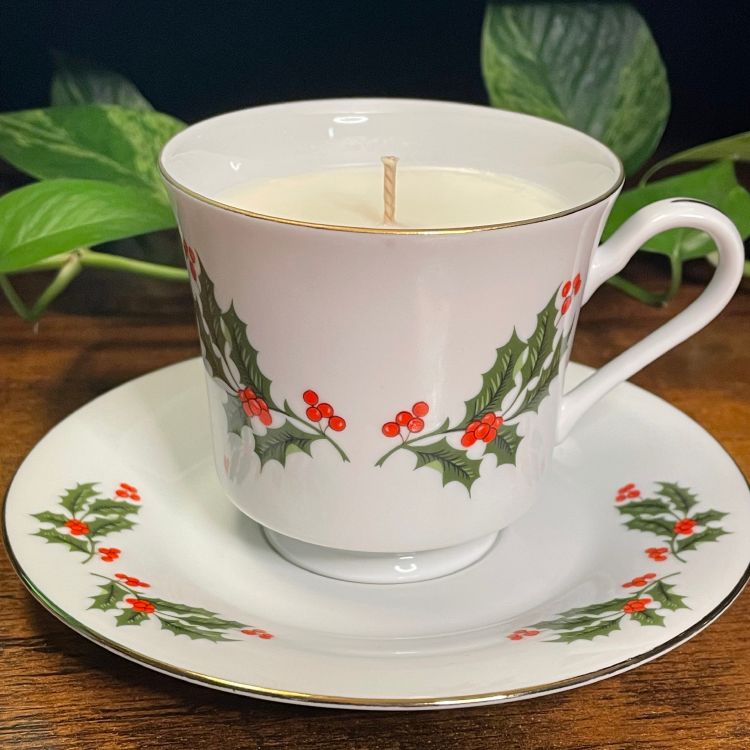 Teacup and saucer with holly design containing a candle, on a wooden surface with green leaves in the background.