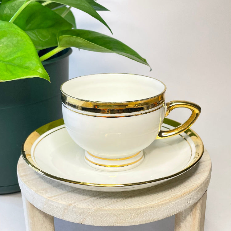 White teacup with gold trim on a matching saucer, placed on a small wooden table with a plant in the background.