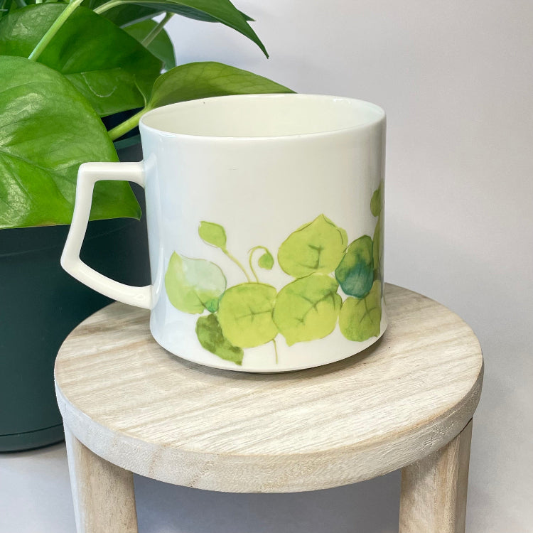 White mug with green leaf design on a wooden stool with a plant in the background
