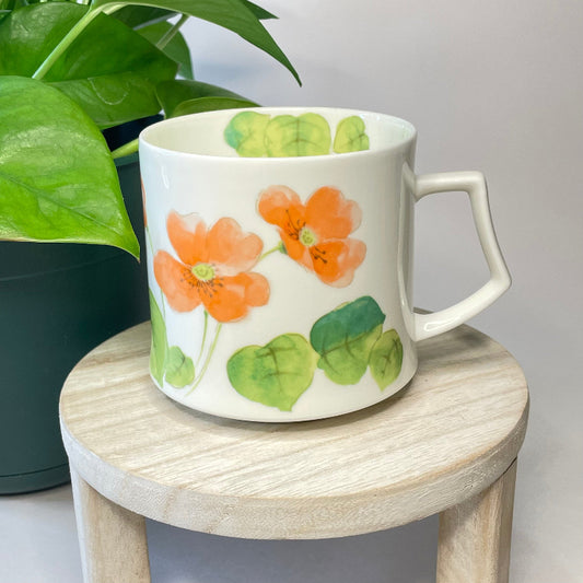White mug with orange flowers and green leaves on a wooden stool with a plant in the background