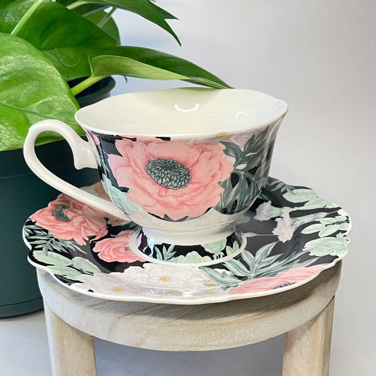 Floral teacup and saucer set on a small wooden stool with a plant in the background.