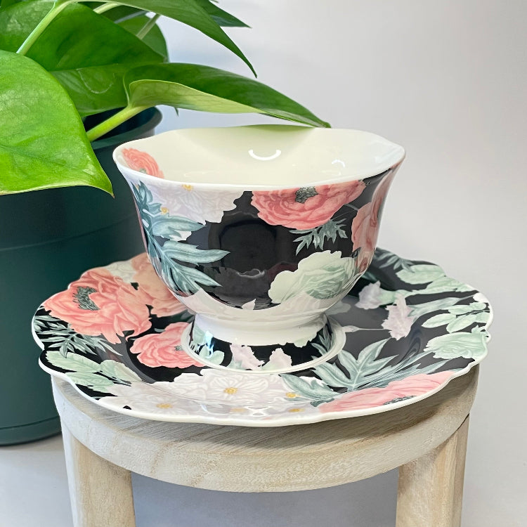 Floral teacup and saucer set on a small wooden stool with a plant in the background.
