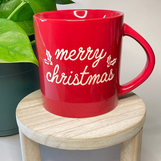 Red mug with 'merry christmas' text on a wooden stool with a plant in the background