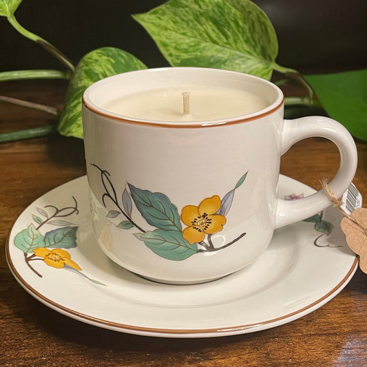 White teacup candle with saucer featuring yellow flowers and green foliage.