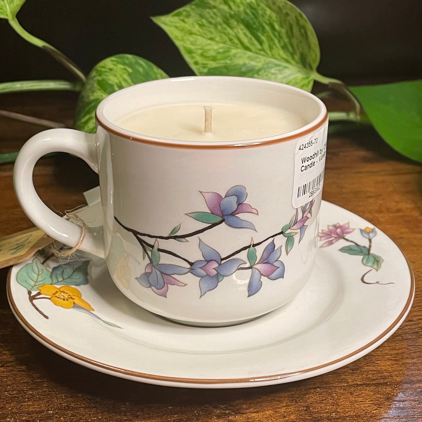 Candle in a decorative teacup with floral design on a saucer, surrounded by green leaves.