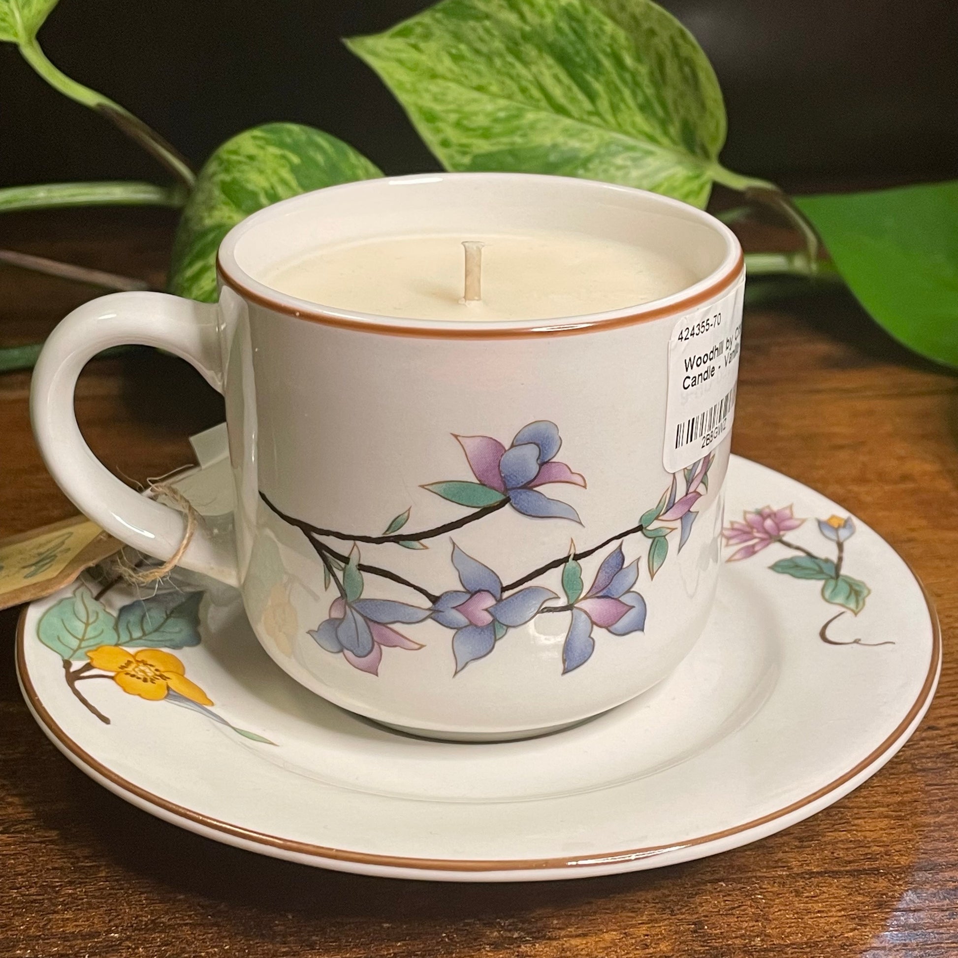 Candle in a decorative teacup with floral design on a saucer, surrounded by green leaves.